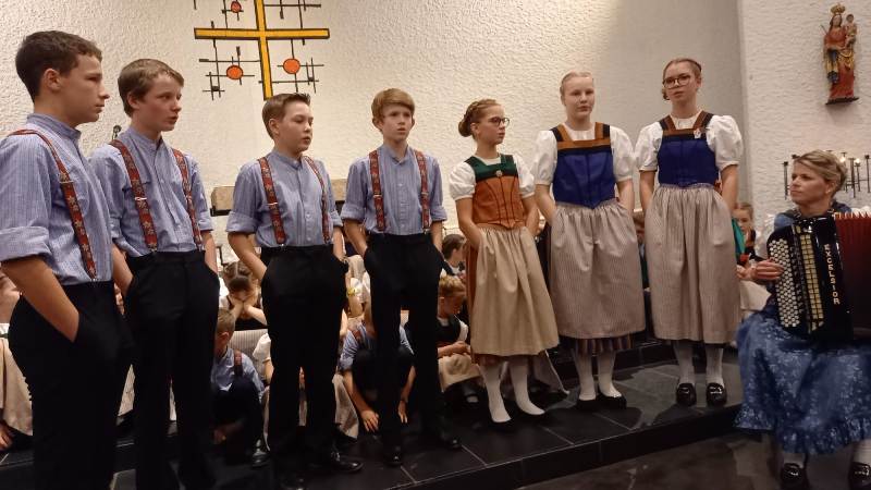 Vier Knaben, drei Mädchen stehen singend 
				nebeneinander, rechts Akkordeonspielerin